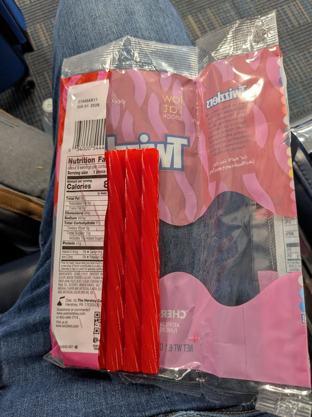 A partially empty Twizzlers candy package rests on someone's denim-clad lap, with several red licorice sticks remaining beside the opened bag. The package's nutrition label and branding are visible.