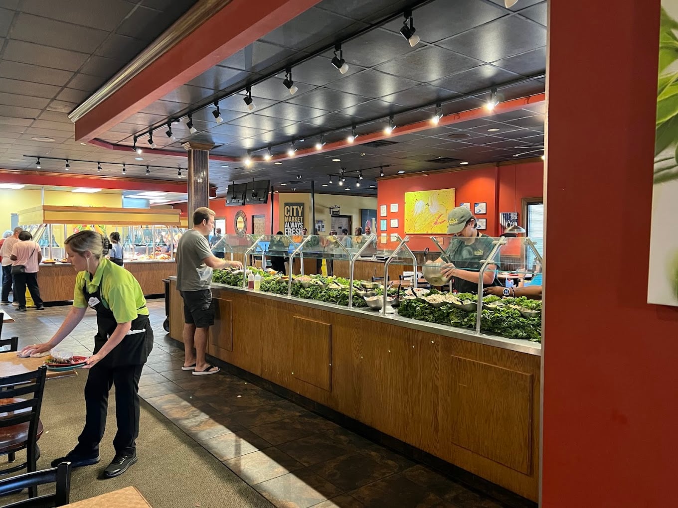 A restaurant interior with people serving themselves at a salad bar. A staff member in a green shirt is cleaning tables in the foreground. The space is brightly lit and has red and yellow walls.