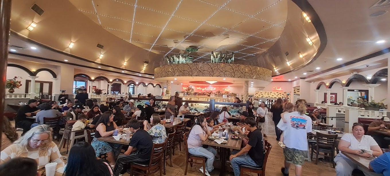 A busy restaurant with many people dining at wooden tables. The ceiling is decorated with lights, and there is a second-floor balcony above the counter. The atmosphere is lively and warm.