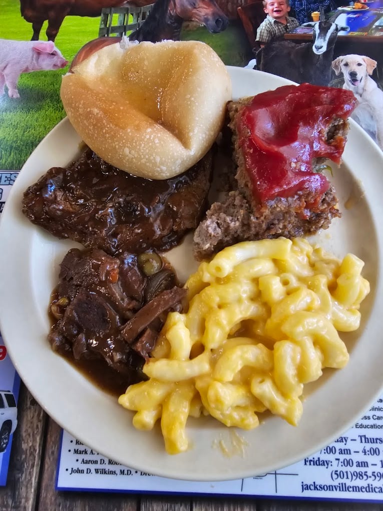 A plate with a bread roll, a slice of meatloaf topped with ketchup, baked macaroni and cheese, and a serving of beef stew with vegetables, set on a colorful placemat.