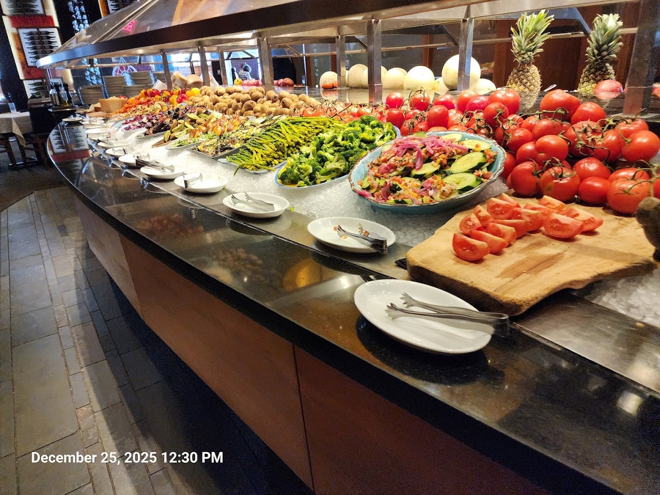 A buffet display with various fresh vegetables, salads, tomatoes, pineapples, and melons on ice, with plates and serving utensils arranged in front. The buffet is indoors with wooden flooring.