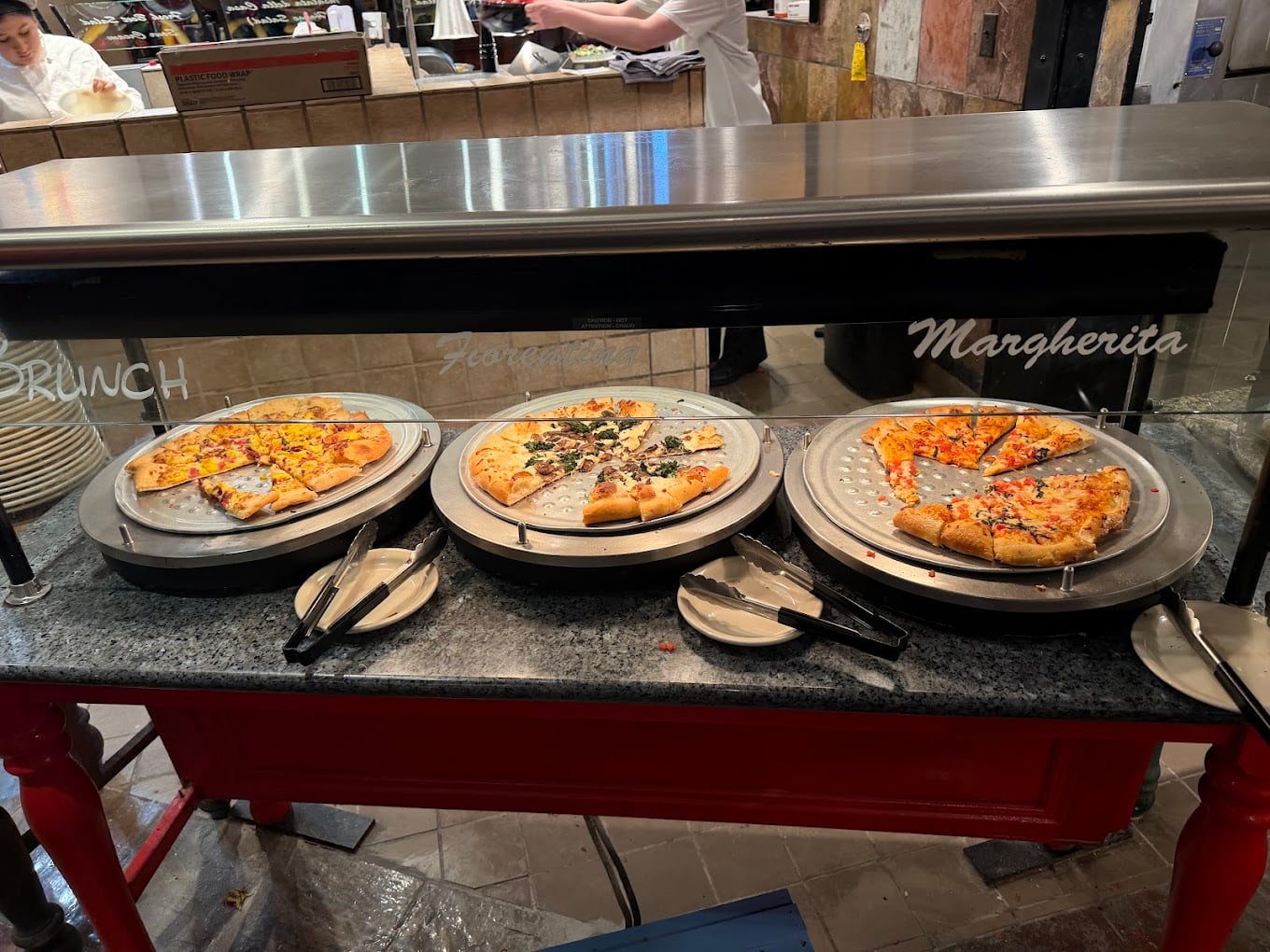 Three pizzas—brunch, vegetarian, and margherita—are displayed on trays under a glass counter with serving tongs and plates in front of each. A person works in the background behind the counter.