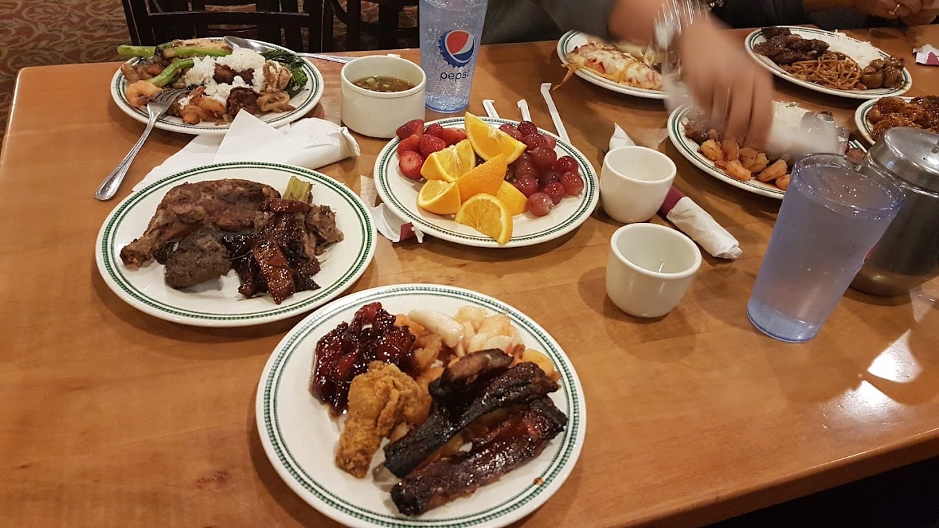 Plates filled with various foods including ribs, steak, fried chicken, fruit, and pasta are set on a wooden table, along with cups, water, a Pepsi, and utensils. Several people are reaching for food.