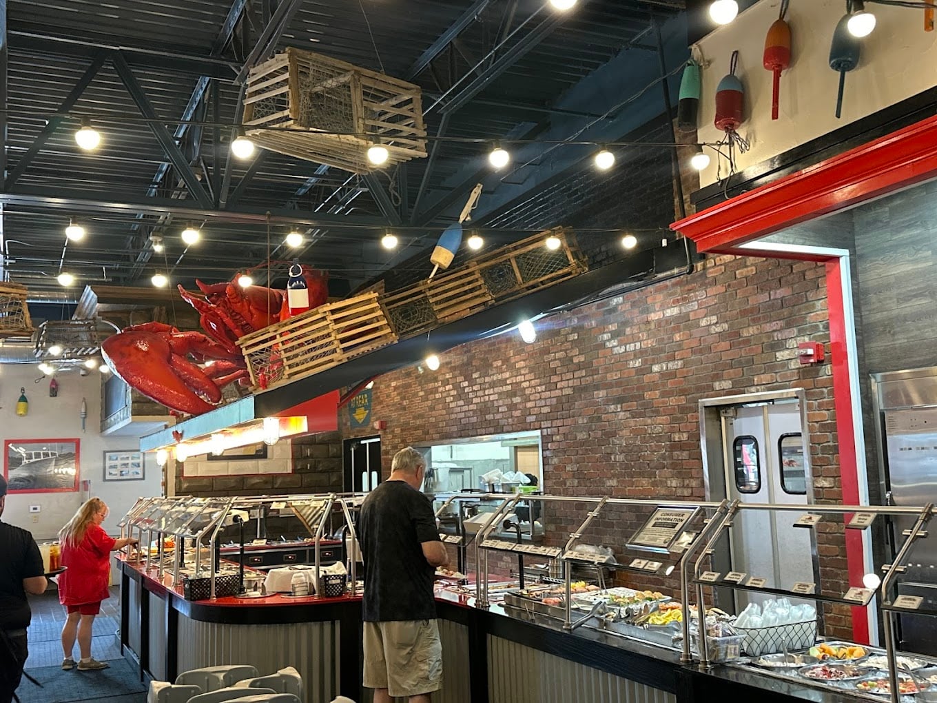 A seafood buffet restaurant with brick walls, fishing decor, and crate and buoy decorations hanging above the food stations. People are serving themselves from the buffet under bright ceiling lights.