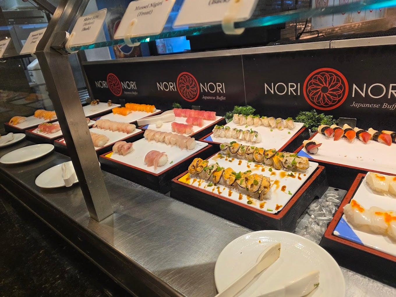 A buffet counter displays various types of sushi and nigiri on white trays, with plates and serving tongs in front. The background features “NORI Japanese Buffet” signs.