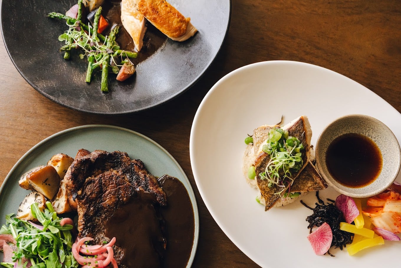Three artfully plated dishes sit on a wooden table: a dark sauced steak with greens and roasted vegetables, a grilled fish fillet with vegetables and dip, and a roasted poultry dish with asparagus and microgreens.