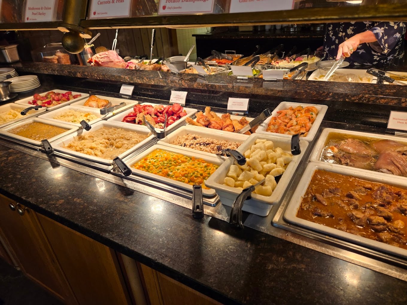 A buffet with trays of various hot foods, including roasted meats, potatoes, carrots, rice, vegetables, and gravy dishes on a dark countertop. A person serves food in the background.