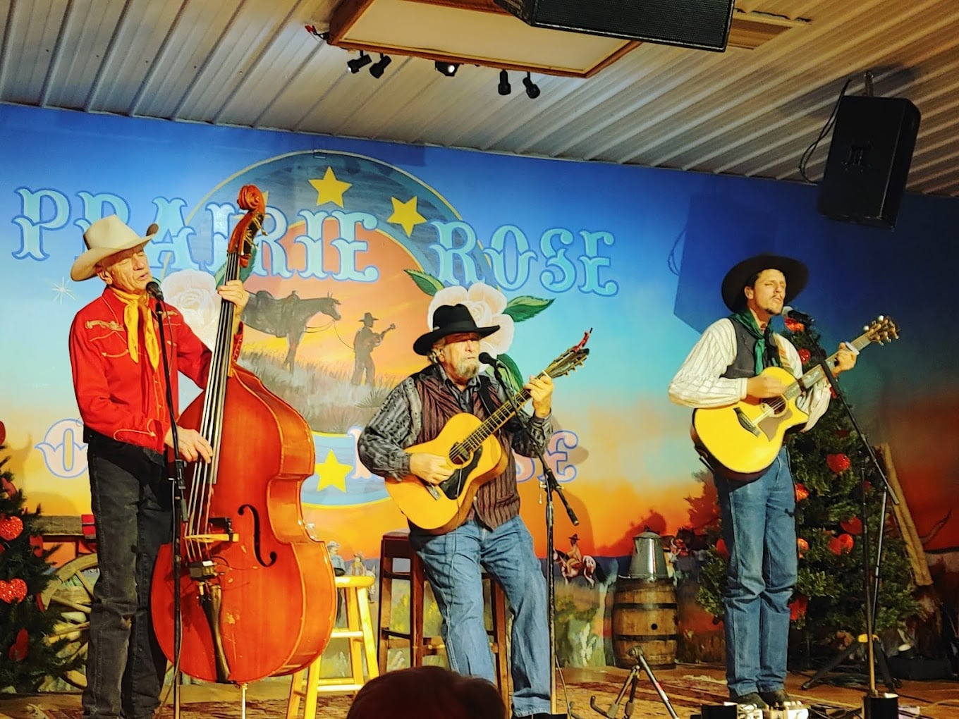 Three musicians dressed in Western attire perform on stage. One plays an upright bass, and the other two play guitars. The backdrop reads "Prairie Rose" with a Western scene and holiday decorations on either side.