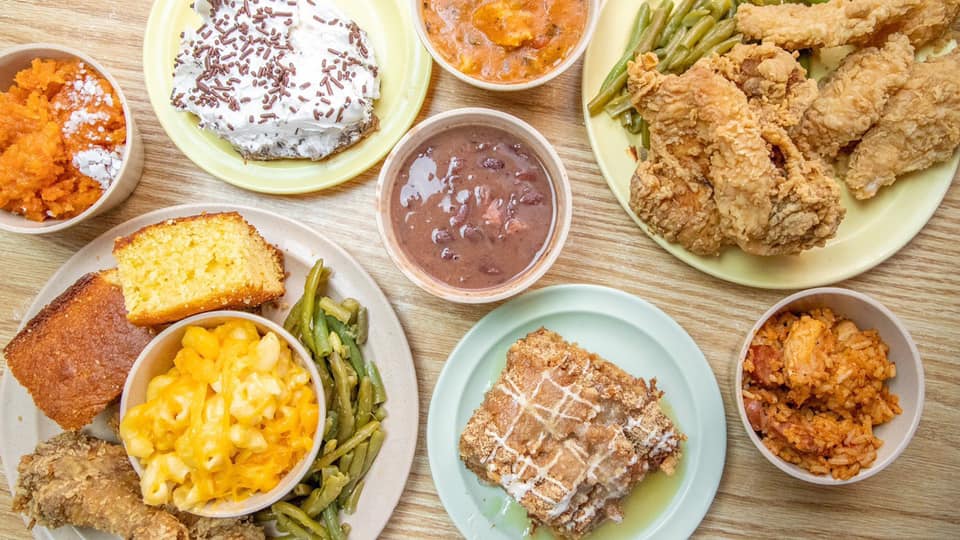 An overhead view of plates and bowls filled with fried chicken, cornbread, macaroni and cheese, green beans, baked beans, rice, sweet potato, and slices of cake on a wooden table.