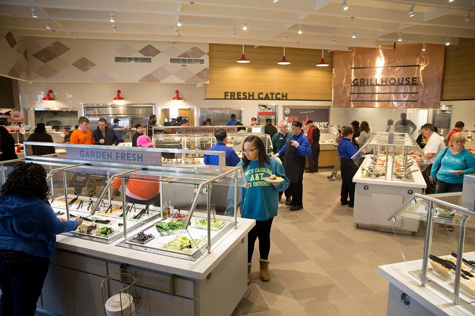 People serve themselves food at various stations in a spacious, well-lit cafeteria with salad, seafood, and grill options. Some diners are selecting items, while others are walking around or waiting in line.