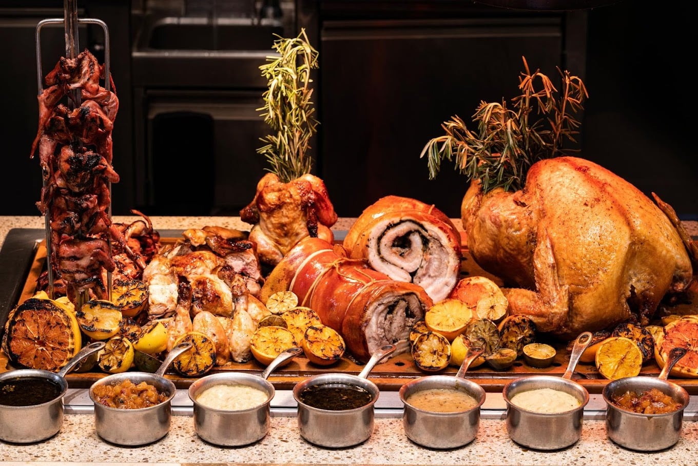 A buffet display featuring roasted meats including turkey, pork, and chicken with herbs, grilled lemons, and eight small sauce pots arranged in front on a countertop.
