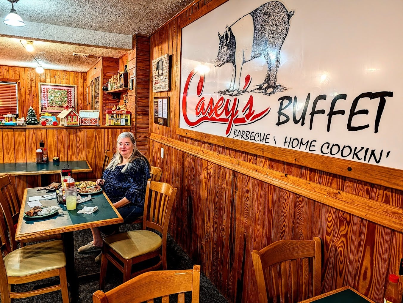 A woman sits at a table with food inside a cozy, wood-paneled restaurant. A large sign on the wall reads "Casey's Buffet Barbecue Home Cookin'" with a drawing of a pig above the text.