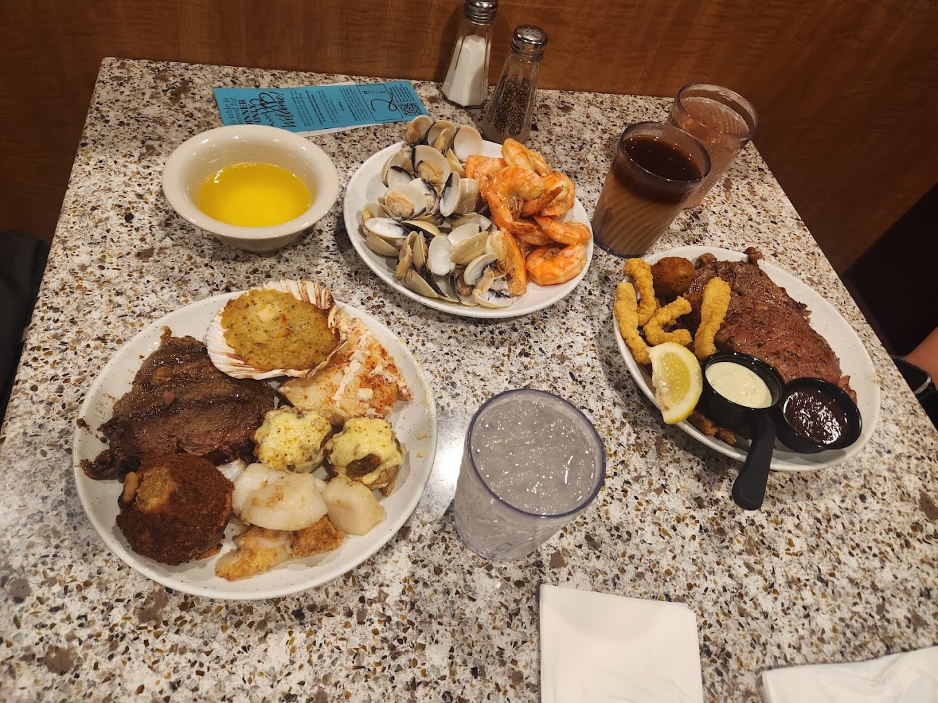 A table with two plates of seafood and steak, a bowl of butter, a plate of clams and shrimp, two drinks, a glass of water, napkins, salt and pepper shakers, and a blue menu on a speckled surface.