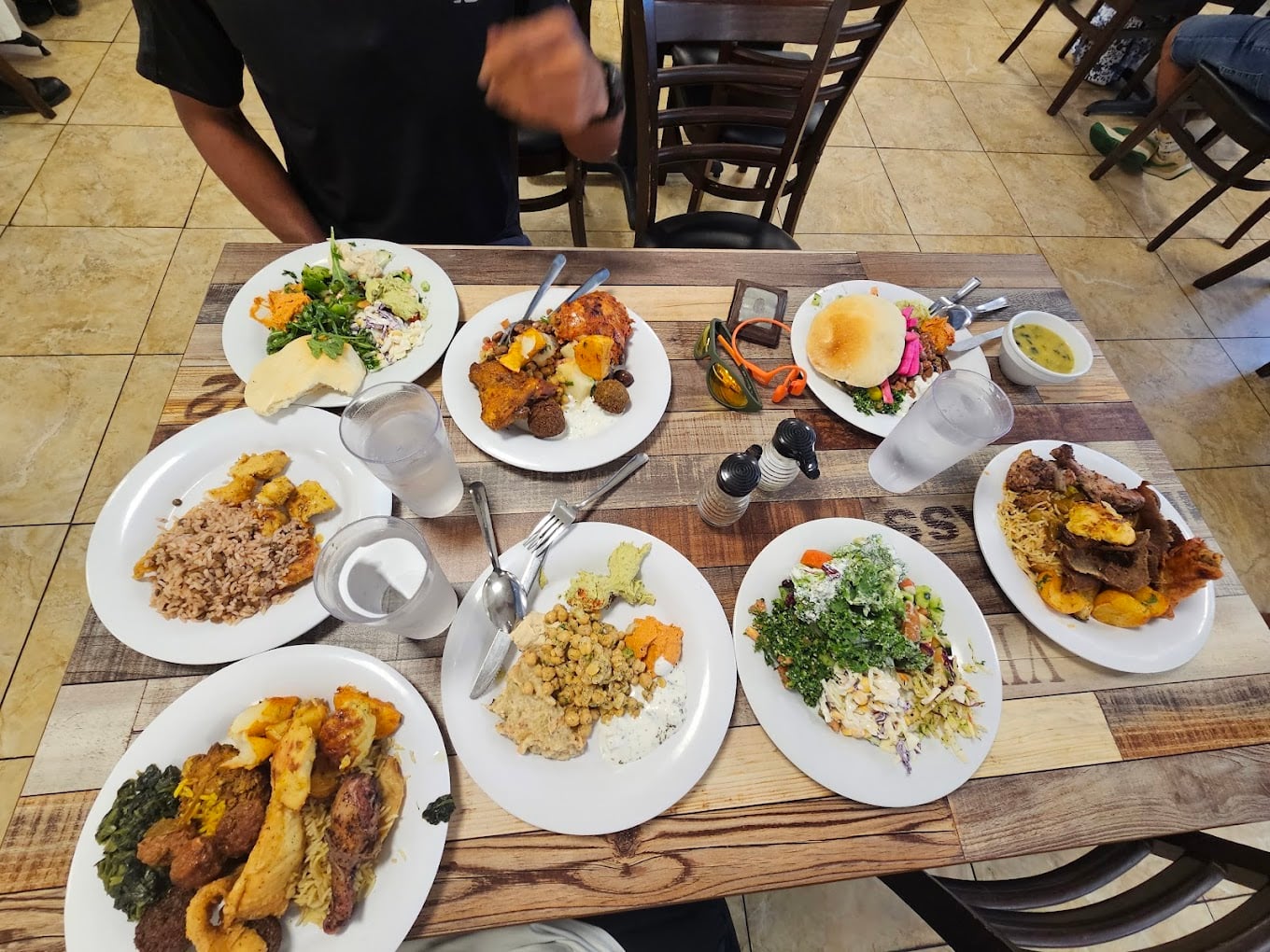 A table set with eight plates of diverse Caribbean dishes, including rice and beans, vegetables, chicken, greens, and salad. Two glasses of water and utensils are also present; a seated person’s arm is visible at the top.