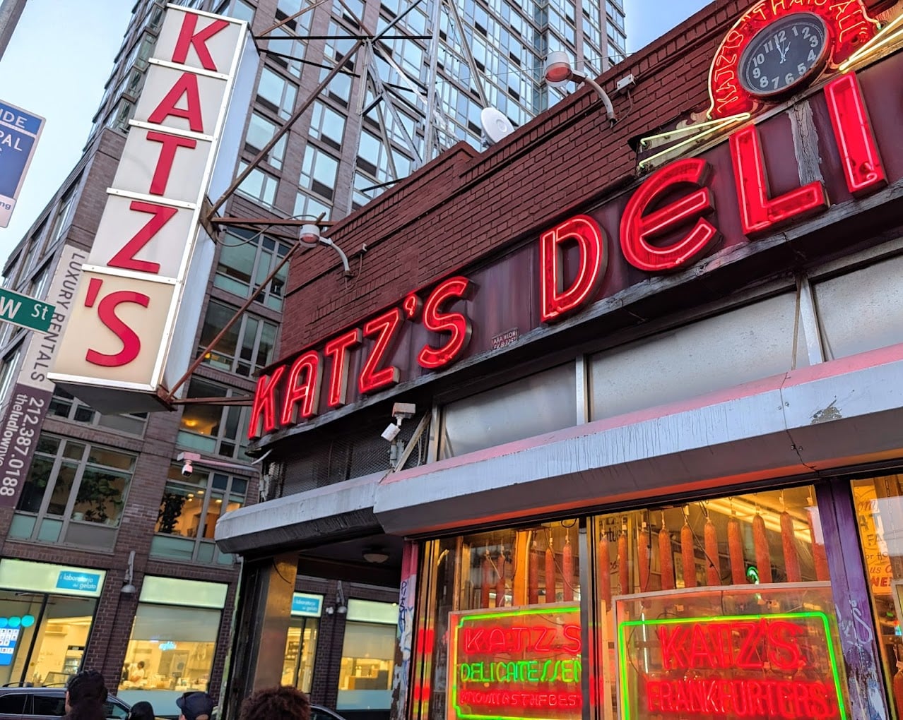 Street view of Katz's Delicatessen in New York City, featuring its large red neon signs and storefront windows, with a clock and tall buildings visible in the background.