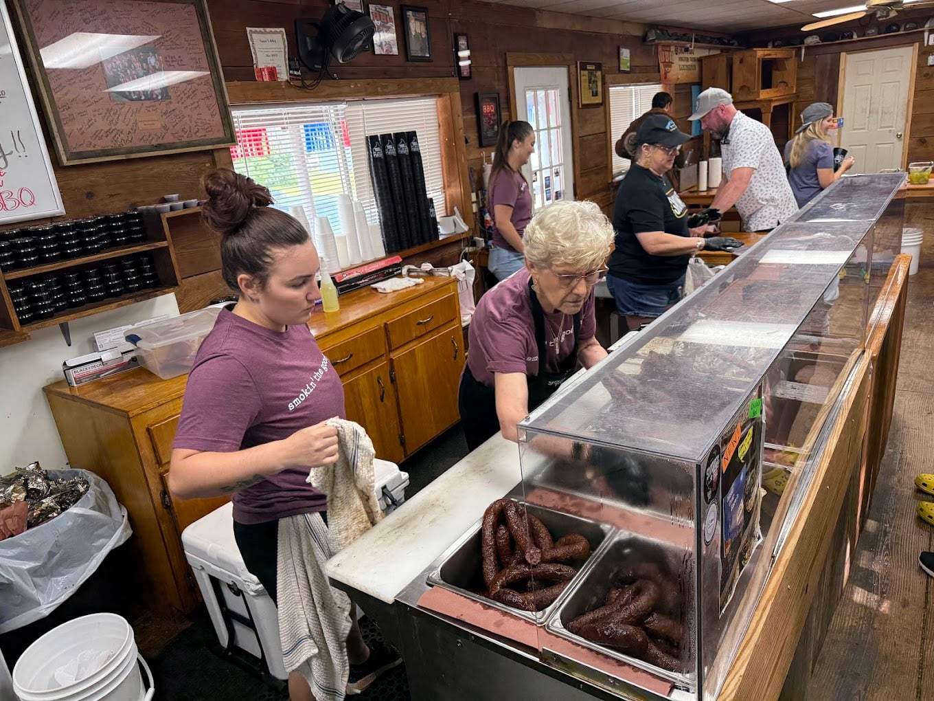 Several people work behind a counter at a rustic deli or BBQ restaurant, serving sausage links from heated trays. The space has wood-paneled walls and various items and signs on shelves and walls.