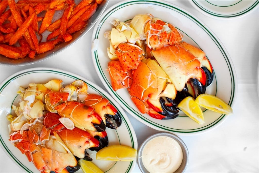 Plates of cracked stone crab claws with lemon wedges and dipping sauce are served on a white tablecloth, alongside a dish of sweet potato fries.