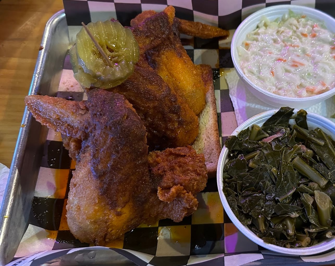 A tray with two pieces of fried chicken topped with pickles, served on white bread, alongside bowls of creamy coleslaw and cooked collard greens on a checkered paper liner.