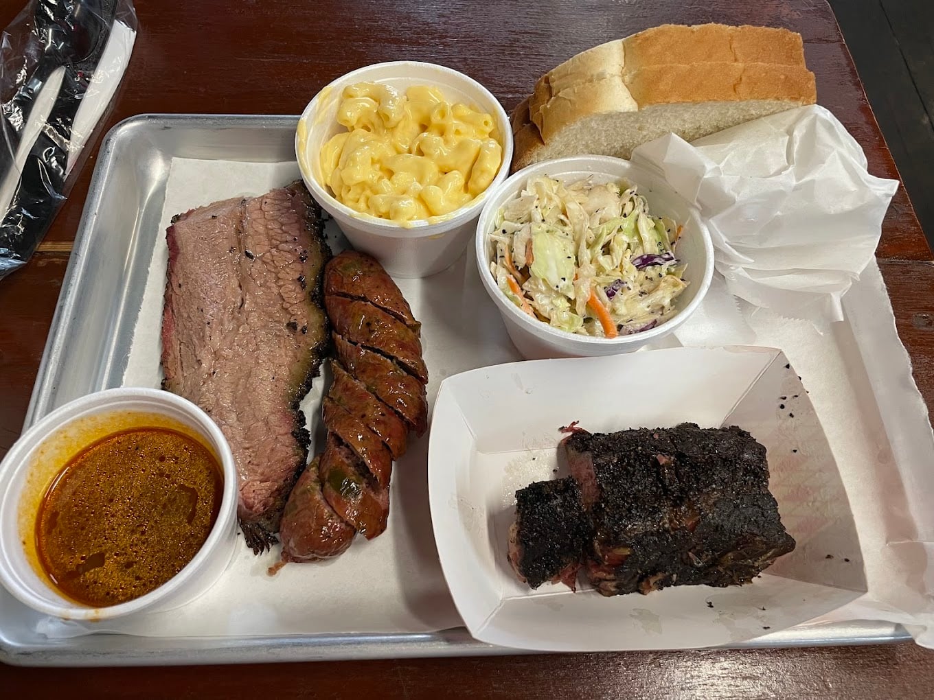 A metal tray with sliced brisket, sausage, a cup of barbecue sauce, macaroni and cheese, coleslaw, bread, and a serving of ribs with a dark crust, all arranged on parchment paper.