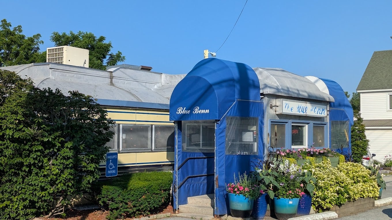 A vintage diner called "Blue Benn" with a blue entrance, surrounded by lush green bushes and colorful potted flowers. A handicap accessible parking sign is visible near the entrance on a sunny day.