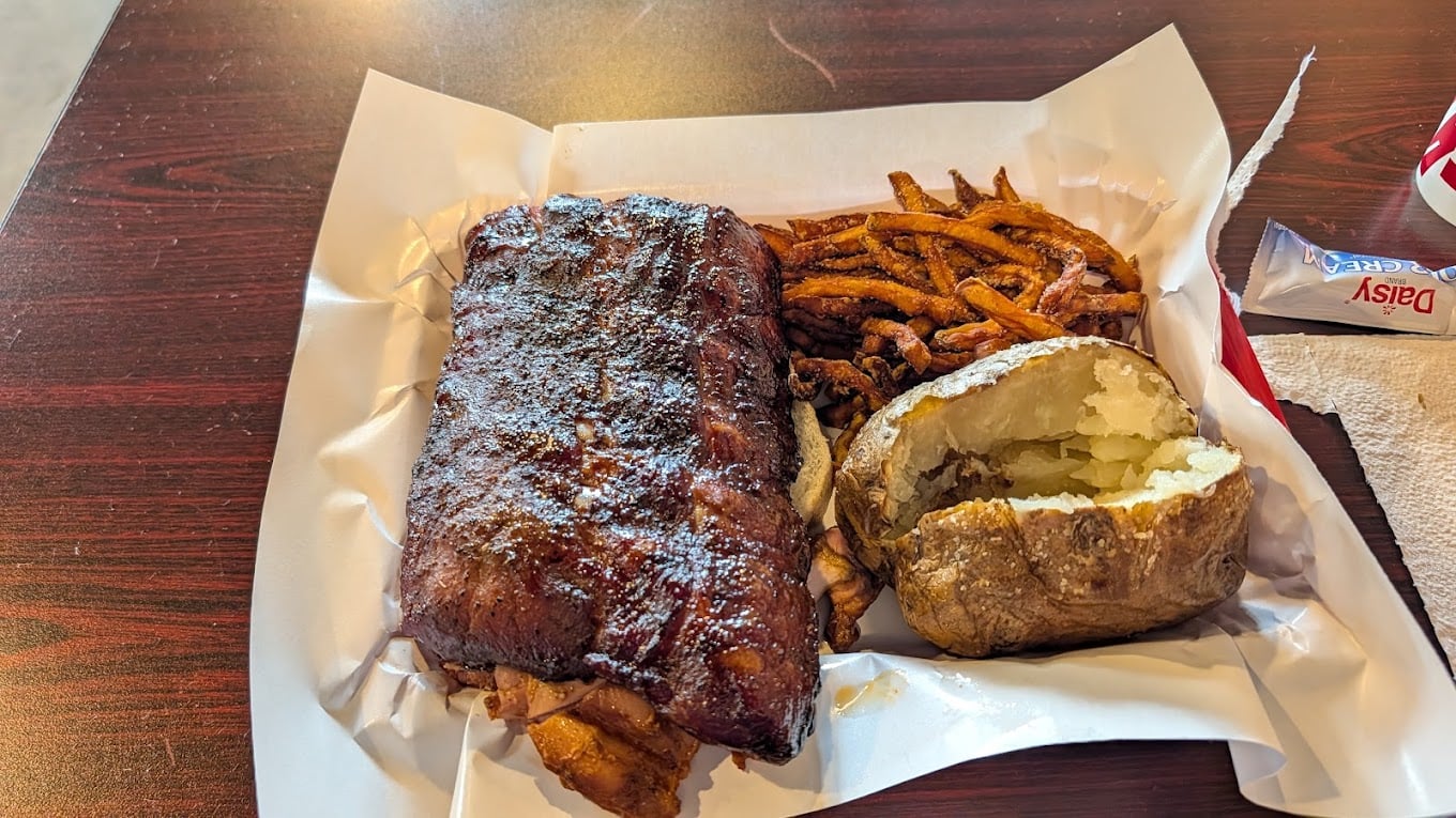 A tray with a rack of barbecued ribs, sweet potato fries, and a baked potato, served on white parchment paper on a wooden table. A napkin and a packet of Daisy sour cream are on the side.