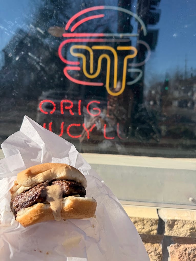 A cheeseburger with melted cheese in the center is held up in front of a window with a neon sign depicting a burger and the words "ORIG JUICY LU". Sunlight shines on the scene.