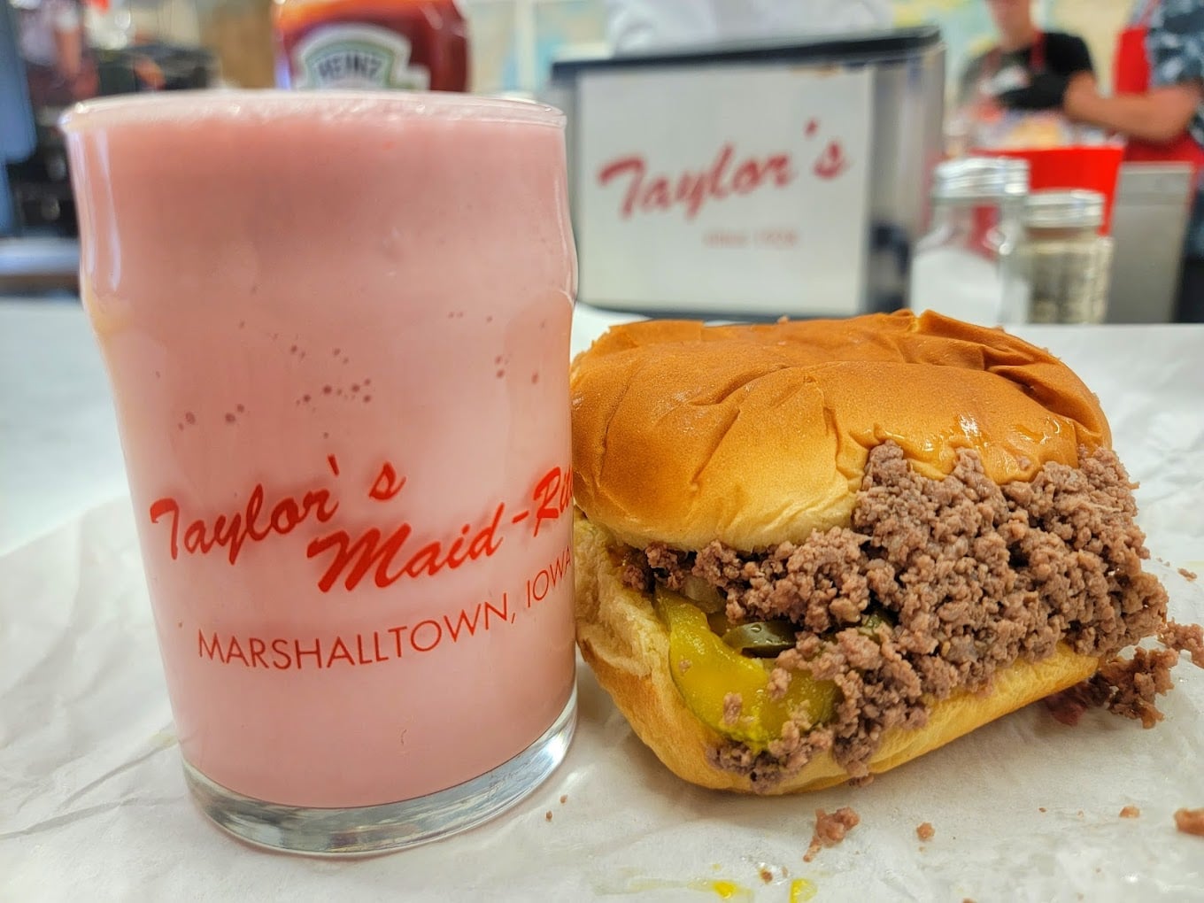 A pink milkshake in a glass labeled "Taylor's Maid-Rite, Marshalltown, Iowa" sits next to a loose meat sandwich with pickles on a soft bun, both on white parchment paper in a diner setting.