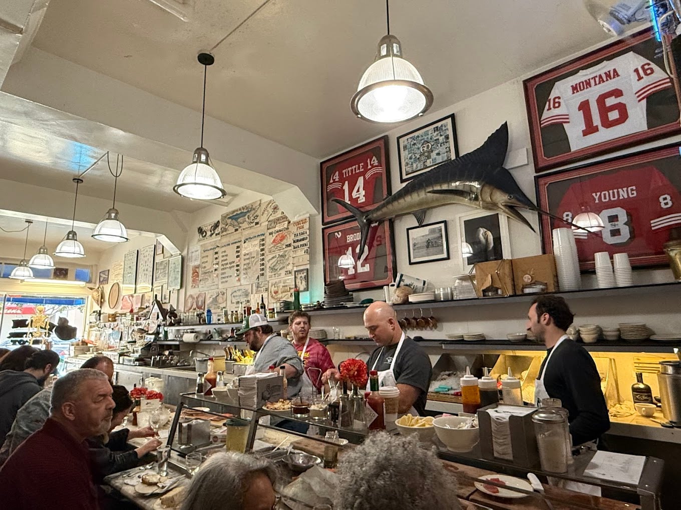 A busy diner interior with people eating at a counter, servers behind it, and sports jerseys, a large mounted fish, and framed photos decorating the walls. Bright overhead lights illuminate the scene.