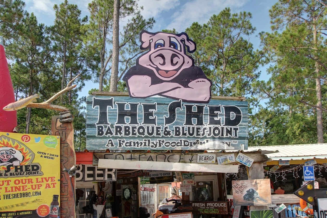 Outdoor BBQ restaurant entrance with a large cartoon pig sign reading "The Shed Barbeque & Blues Joint, A Family Food Drinkery." Surrounding signs and decorations, with trees and sky in the background.