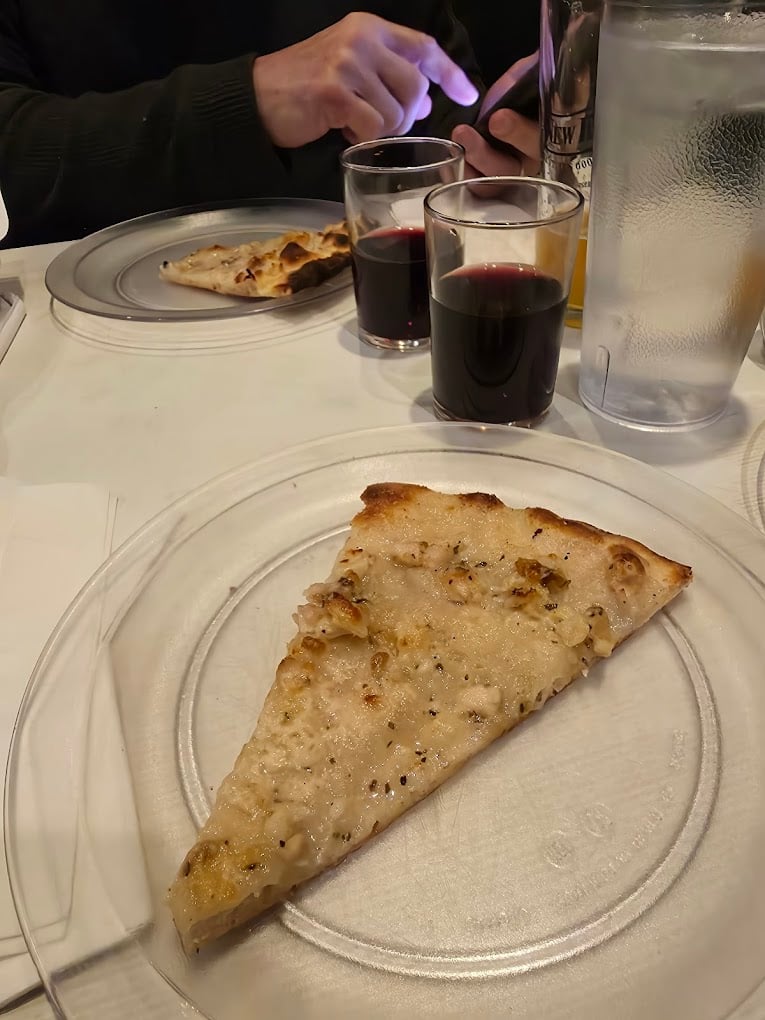 A slice of white pizza on a clear plate sits on a table with a glass of red wine, a glass of water, and another plate. In the background, a person uses a smartphone near another plate and glass of wine.