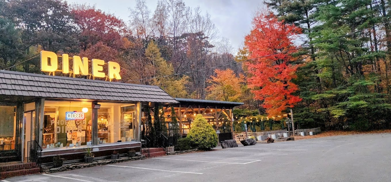 A roadside diner with a large yellow "DINER" sign is closed at dusk, surrounded by trees with colorful autumn foliage and an empty parking lot. String lights decorate the outdoor seating area.