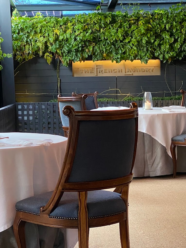 A dining area at the famous restaurant The French Laundry features elegant wood chairs with gray upholstery, white tablecloths, and a green ivy-covered wall displaying the restaurant's name.