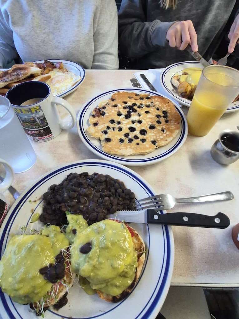 Two people sit at a table with plates of breakfast food, including blueberry pancakes, eggs Benedict with avocado sauce and black beans, and a plate of eggs and toast. There are also mugs, a glass of orange juice, and utensils.