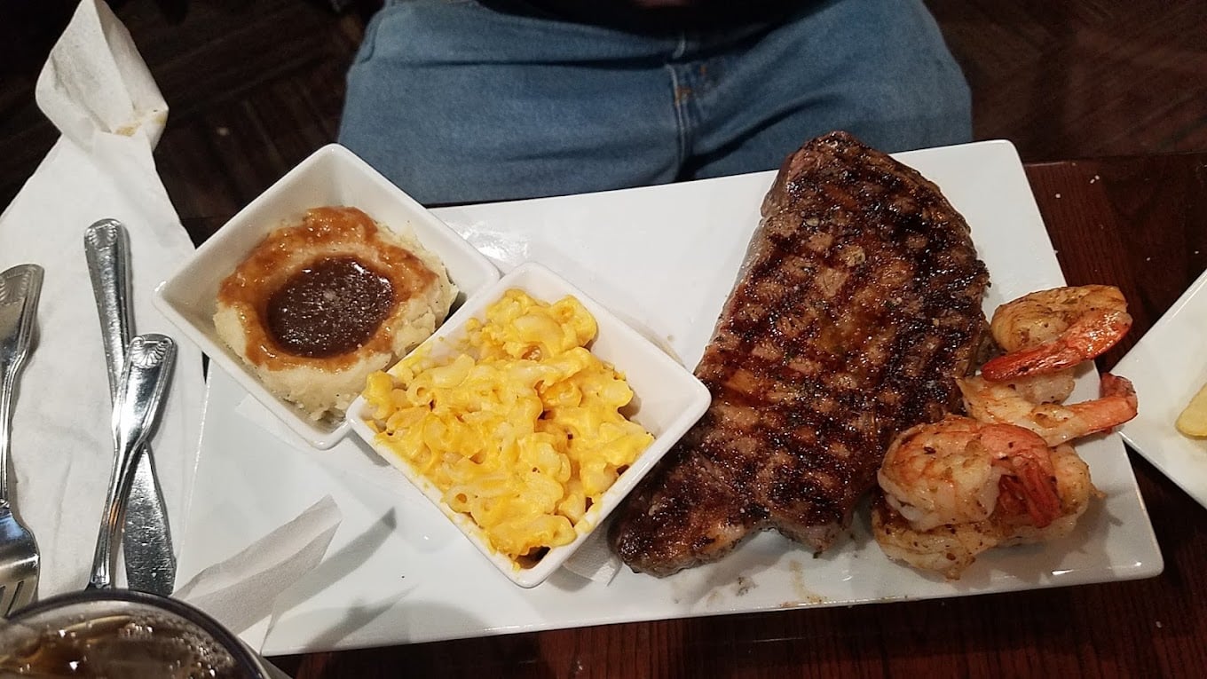 A rectangular plate with a grilled steak, three grilled shrimp, mashed potatoes with brown gravy, and macaroni and cheese in small square bowls, with utensils and a napkin beside the plate.