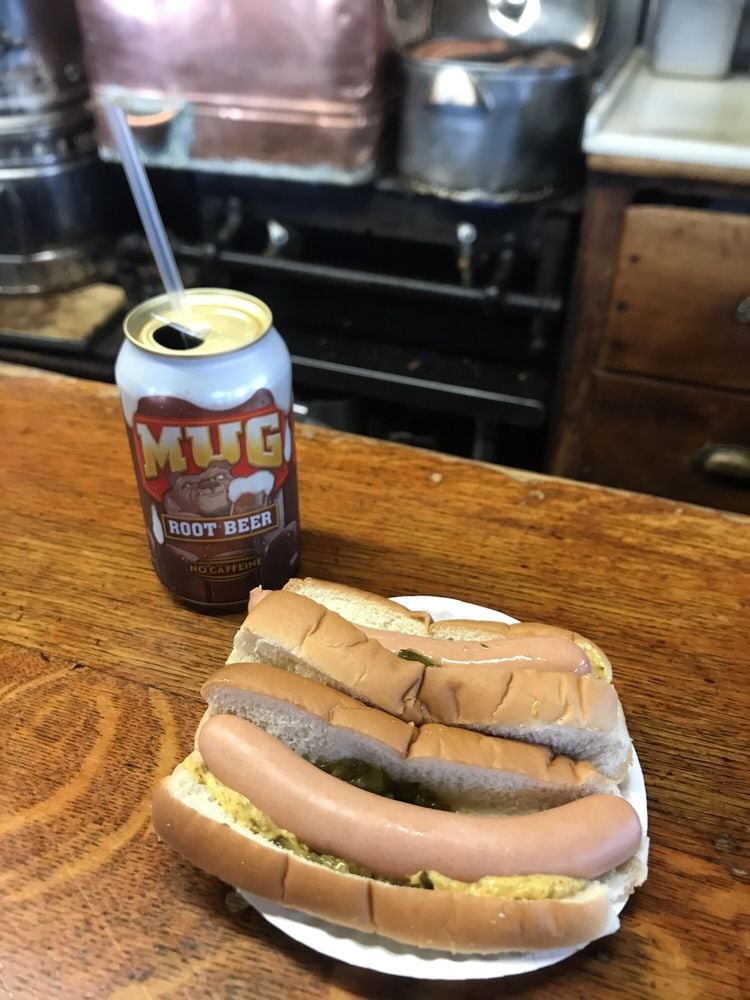 A can of Mug Root Beer with a straw and a plate holding three plain hot dogs in buns sit on a wooden counter, with a kitchen scene visible in the background.
