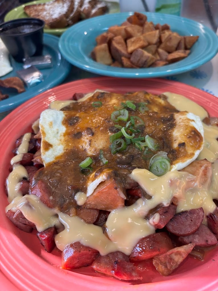 A close-up of a breakfast plate with sausage, diced potatoes, cheese sauce, salsa, and a fried egg, garnished with green onions. In the background are more diced potatoes and toast with powdered sugar.