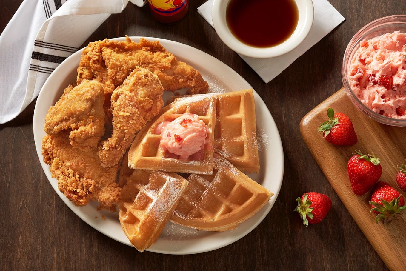 A plate with crispy fried chicken, golden waffles topped with pink strawberry butter, and a dusting of powdered sugar. A bowl of syrup, fresh strawberries, and a dish of strawberry butter are on the side.
