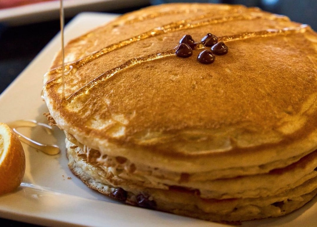 A stack of fluffy pancakes topped with a few chocolate chips and drizzled with syrup on a white plate, served diner-style with a slice of orange on the side—classic cheap diner portions at their best.
