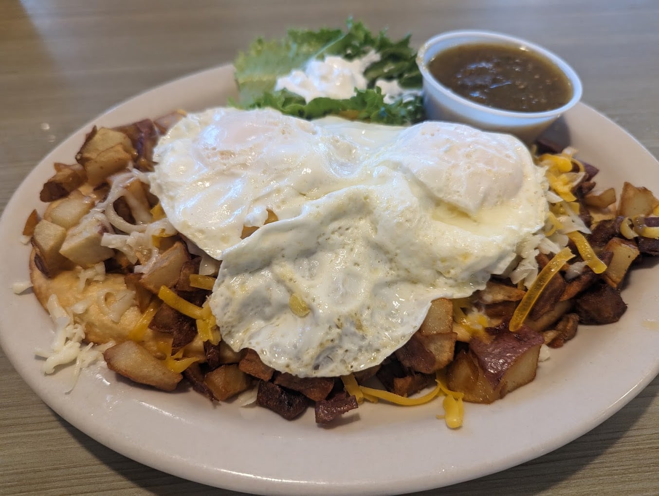 A plate of breakfast hash with diced potatoes, melted cheese, and two fried eggs—served up in cheap diner portions with a side of lettuce, a dollop of sour cream, and a small cup of brown sauce.