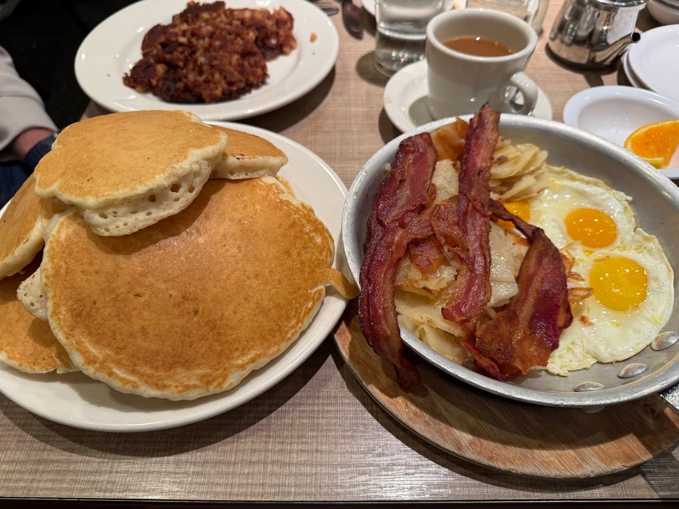 A breakfast spread with a plate of pancakes, a skillet containing sunny-side-up eggs, bacon, and hash browns, a plate of hash, a cup of coffee, and a glass of water on a wooden table—classic cheap diner portions served fresh.