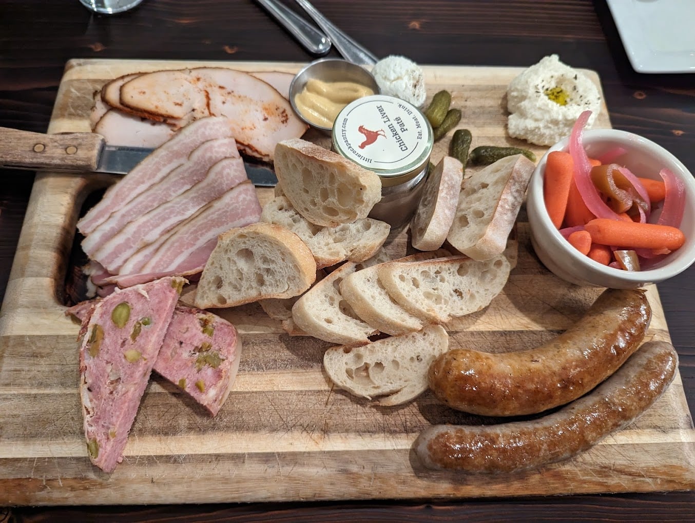 A charcuterie board with sliced cured meats, pâté, sausage links, baguette slices, pickles, mustard, white cheese spreads, and pickled vegetables on a wooden surface—plenty more generous than typical Cheap Diner Portions.