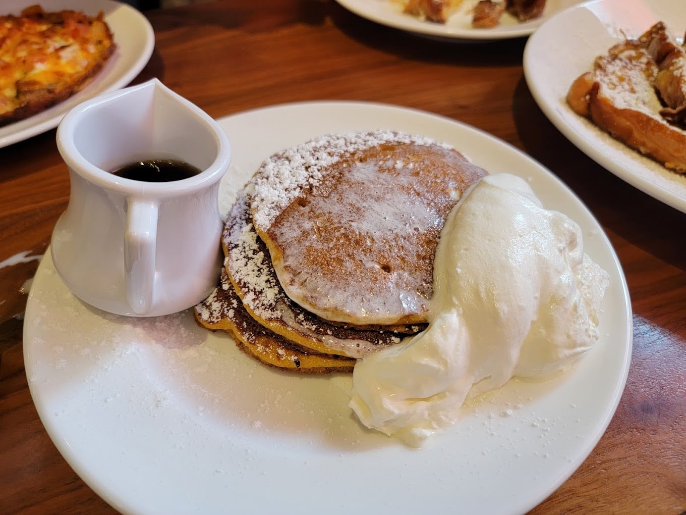 A plate of pancakes topped with powdered sugar, served with a dollop of whipped cream and a small pitcher of syrup on the side, offers classic cheap diner portions on a wooden table.