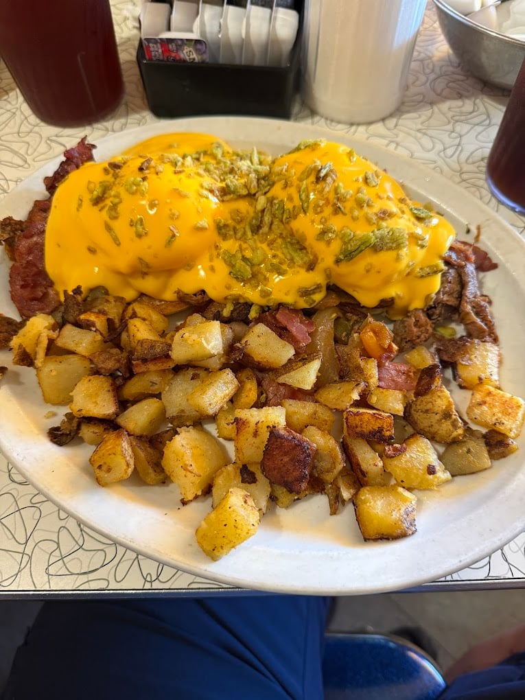 A plate of breakfast potatoes and a large serving of eggs covered in melted cheddar cheese and green herbs, with pieces of bacon underneath—classic cheap diner portions. Condiment containers and drinks are visible in the background.