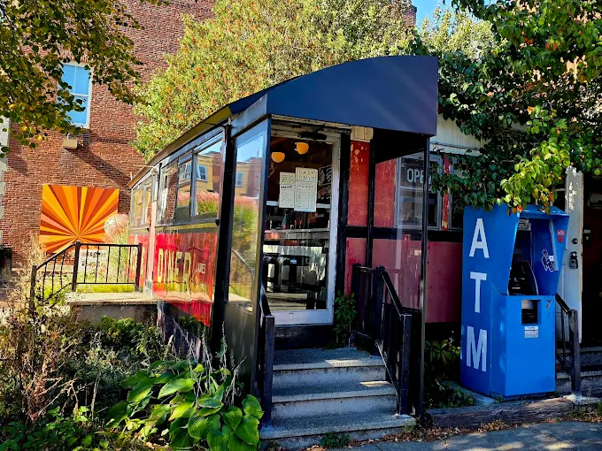 A small roadside restaurant with a glass entrance and a sign on the door sits beside a blue outdoor ATM. Green plants border the walkway, and a vibrant orange geometric mural brightens the nearby brick wall.