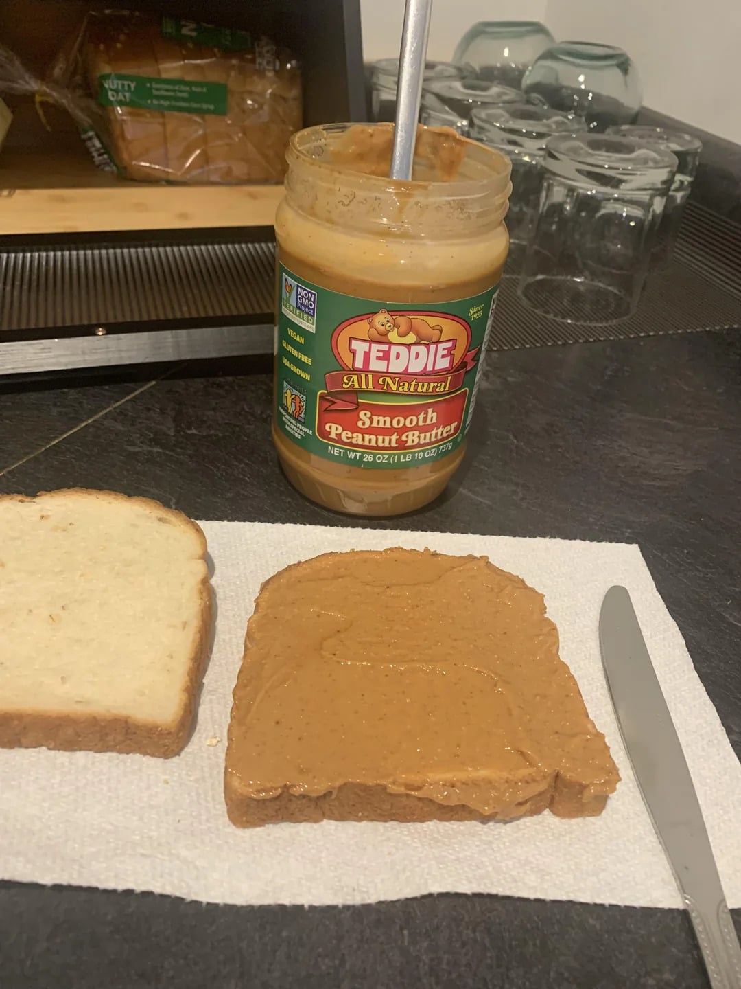 A jar of Teddie Smooth Peanut Butter—one of the best peanut butter brands—sits on a counter with a spoon inside, beside a slice of bread spread with peanut butter, another plain slice, and a knife on a paper towel.