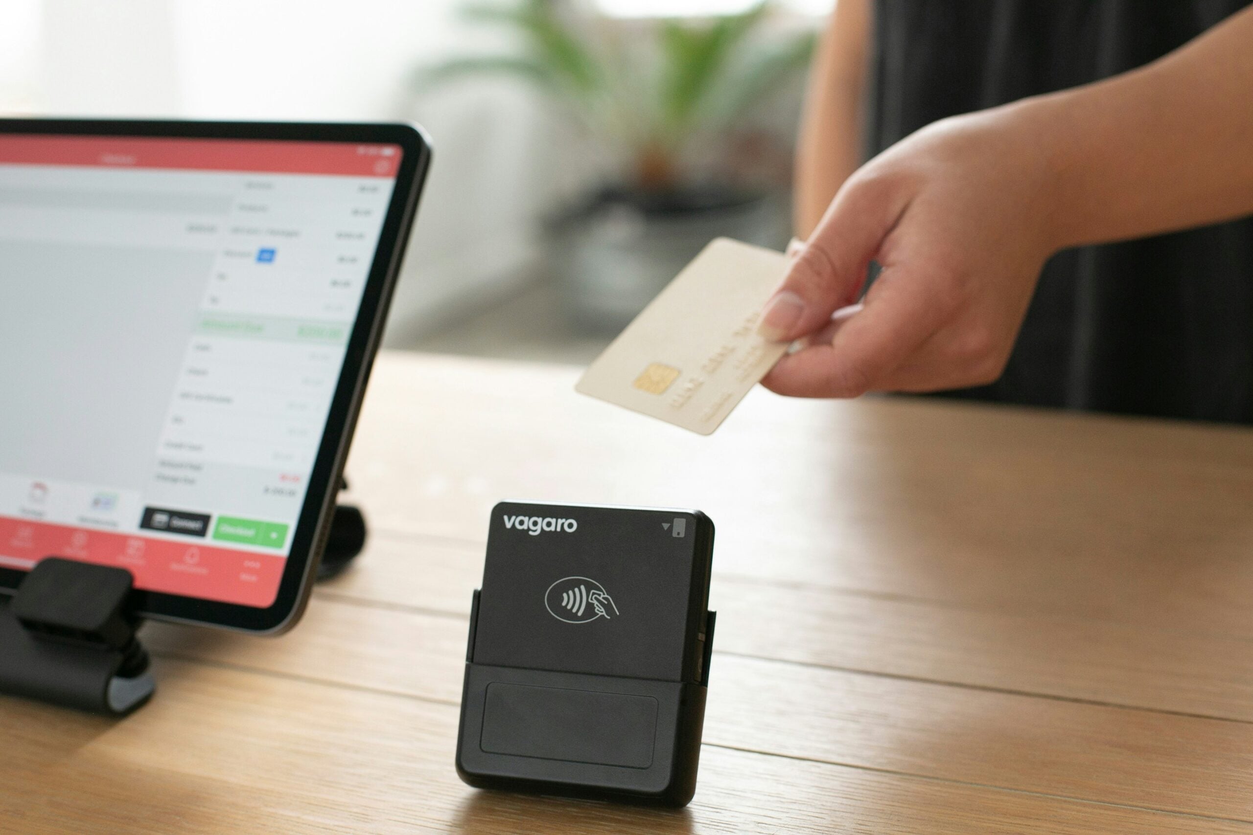 A person holds a credit card above a contactless card reader near a tablet on a wooden counter, preparing to make a payment.
