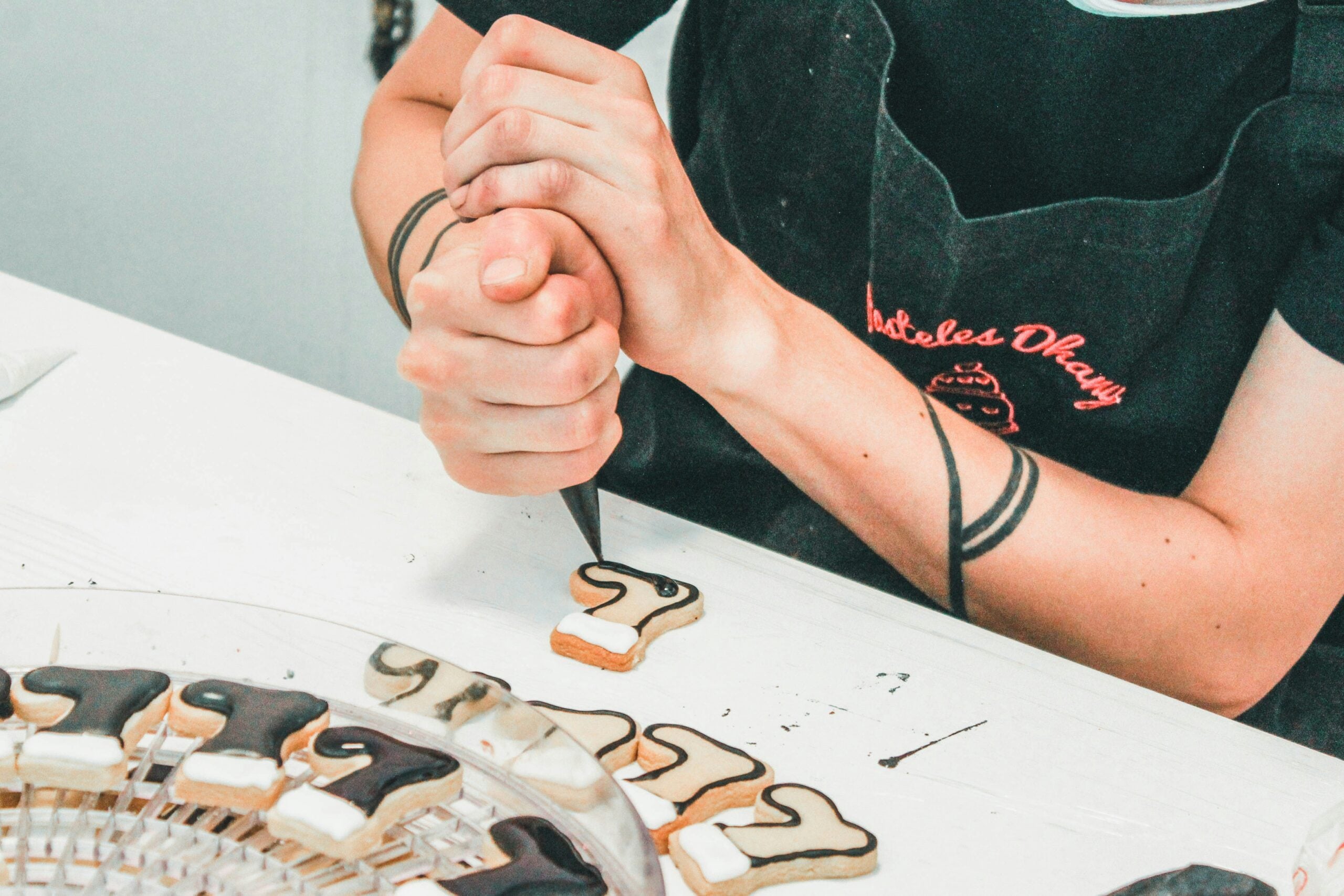 A person wearing a black apron is decorating number-shaped cookies with icing at a table. Some cookies are already decorated with black and white icing and are arranged on a clear tray.