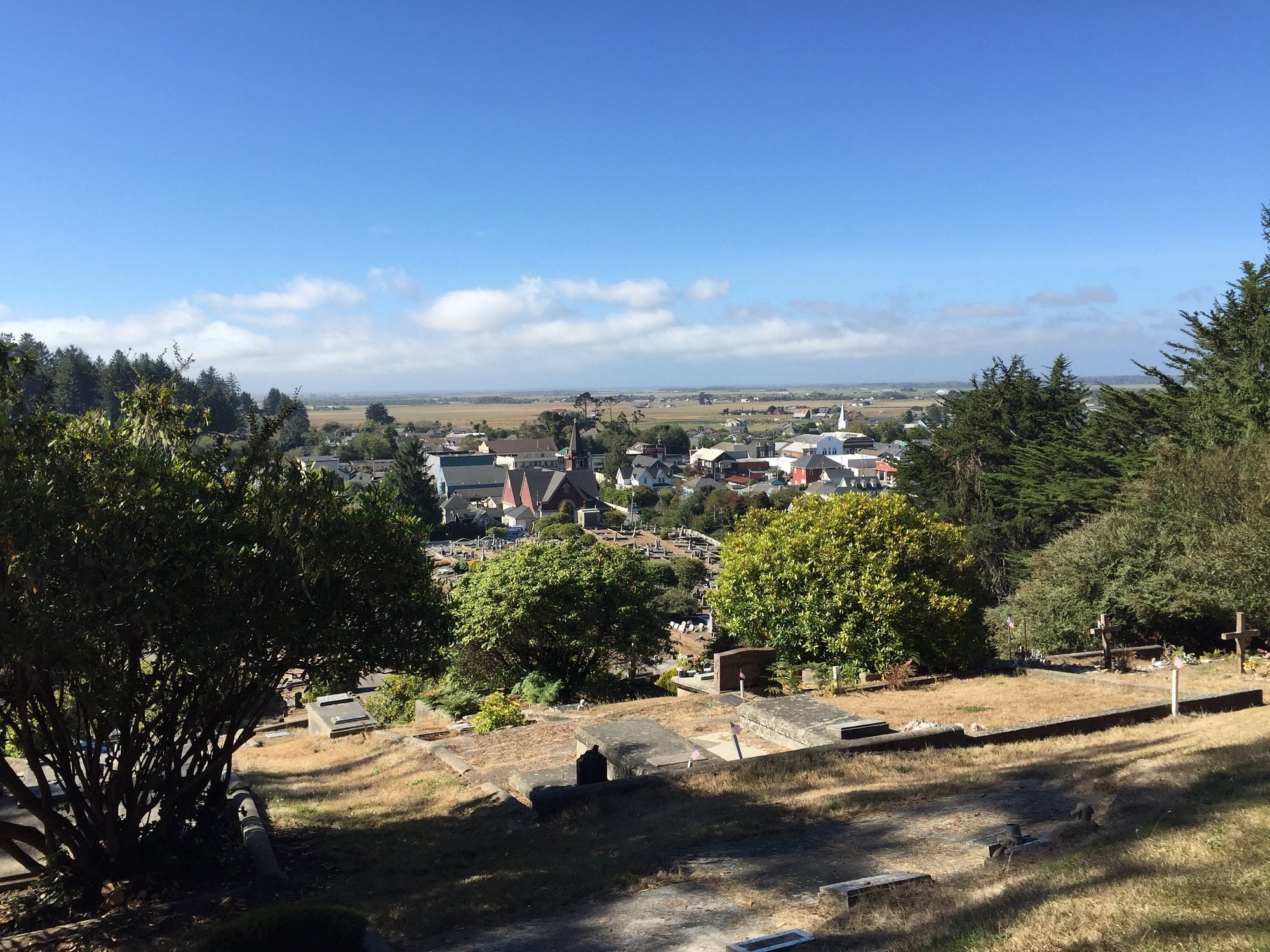A hillside cemetery with gravestones and trees overlooks one of those unique American towns, where a church and houses nestle under a blue sky dotted with scattered clouds. Flat open land stretches in the background.