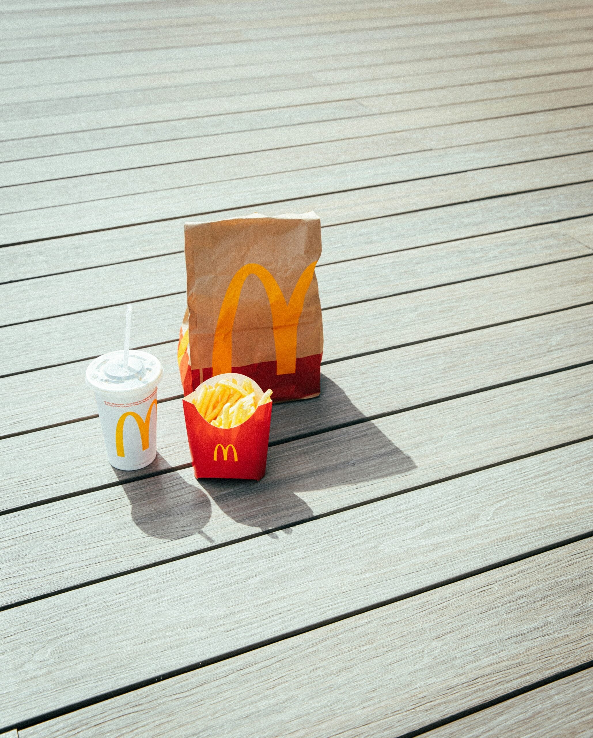 Chain restaurant prices. A McDonald’s meal with a paper bag, fries in a red carton, and a drink cup with a straw sits on a wooden surface in sunlight.