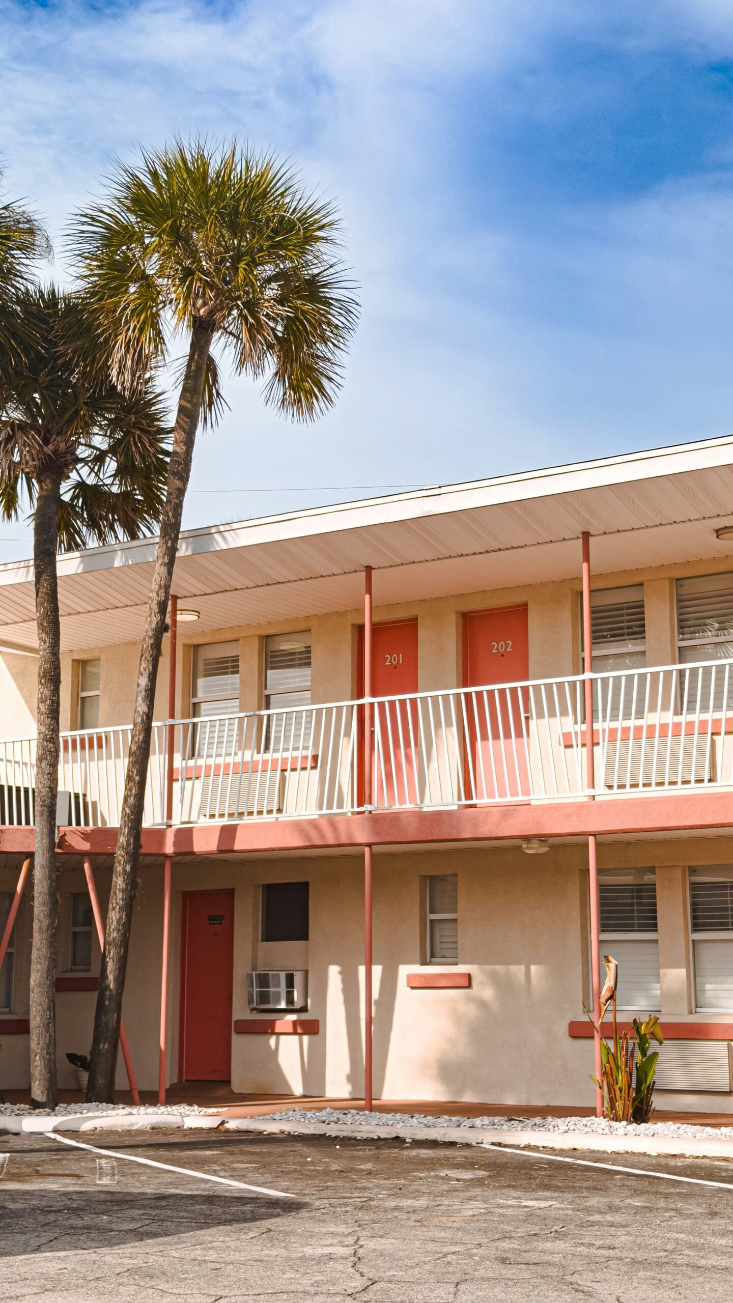 A two-story motel with cream walls, coral-colored doors and railings, and two palm trees in front. The motel has rooms numbered 201 and 202 on the upper floor. The sky is partly cloudy.